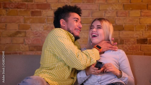 Young african guy strangles his blonde caucasian girlfriend watching TV in cozy home atmosphere.