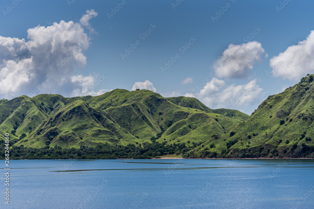 Komodo Island, Indonesia - February 24, 2019: Green mountain range descending on sand beach under blue sky with cloudscape, part of Komodo National Park.