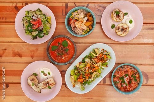 Top view of various vegetarian meals, served on a light wooden table: poke bowl, gazpacho, vegetable salads, BBQ fruits and vegan noodle soup