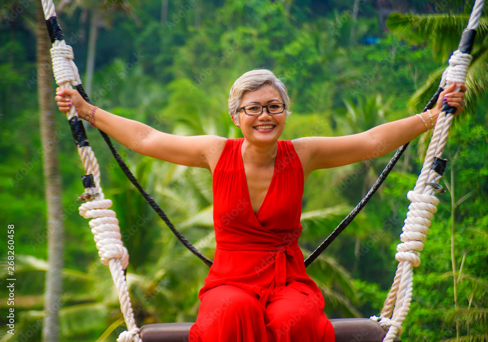 natural lifestyle portrait of attractive happy middle aged 40s - 50s Asian woman with grey hair and stylish red dress riding rainforest swing carefree swinging