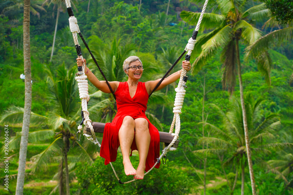 attractive happy middle aged 40s or 50s Asian Indonesian woman with grey hair riding rainforest swing carefree swinging and enjoying tropical jungle adventure