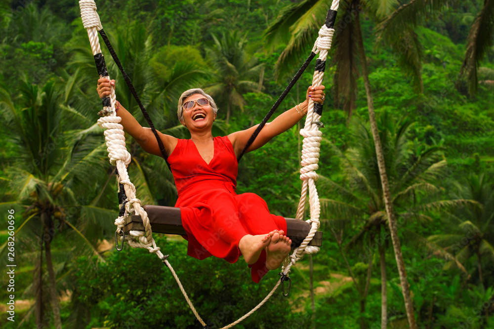 attractive happy middle aged 40s or 50s Asian Indonesian woman with grey hair riding rainforest swing carefree swinging and enjoying tropical jungle adventure