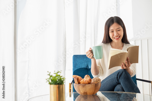 Young beautiful Asian woman relaxing in living room at home, reading books, drinking coffee in the morning with happy feeling