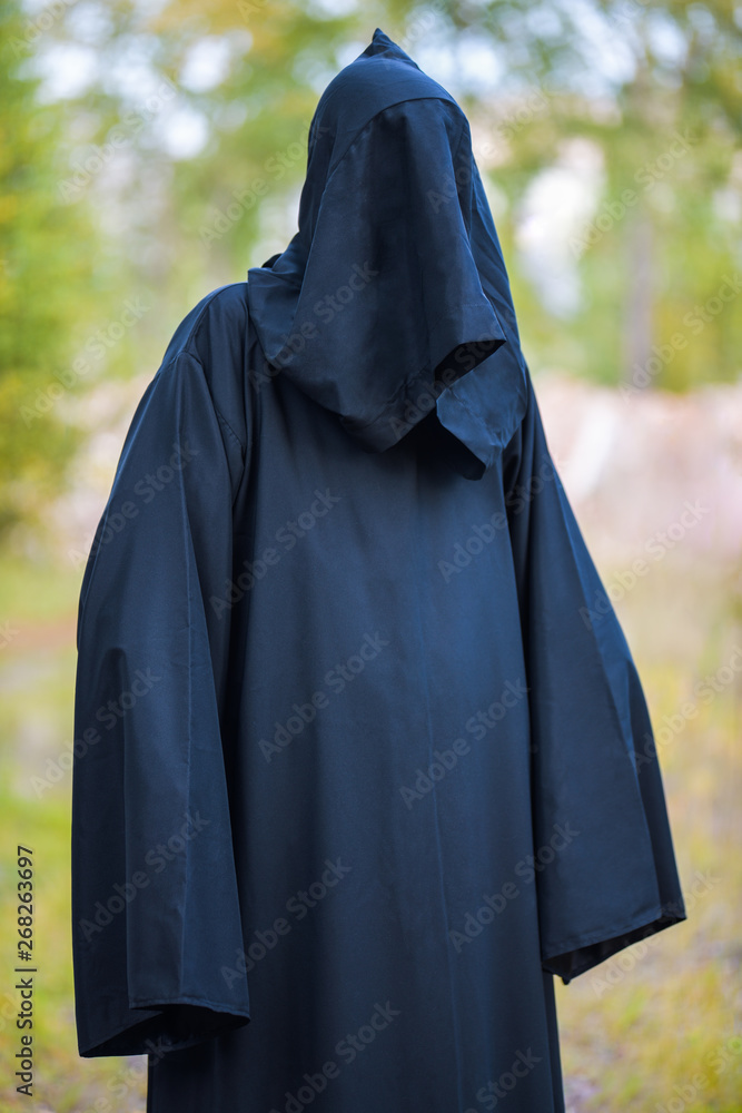 A man in a black suit with a hood stands in the woods on a green background. Vertical photography
