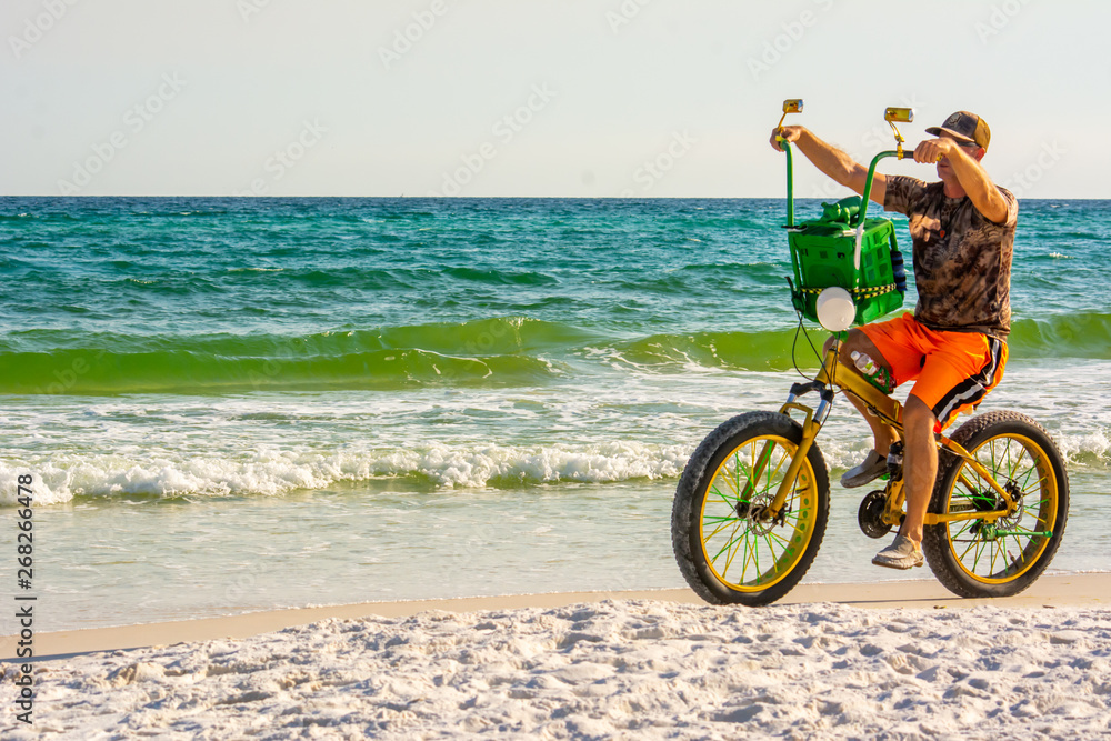 Fototapeta premium Man Riding a Bycicle on White Sand on Blur Ocean Water Background. Destin Beach, Florida