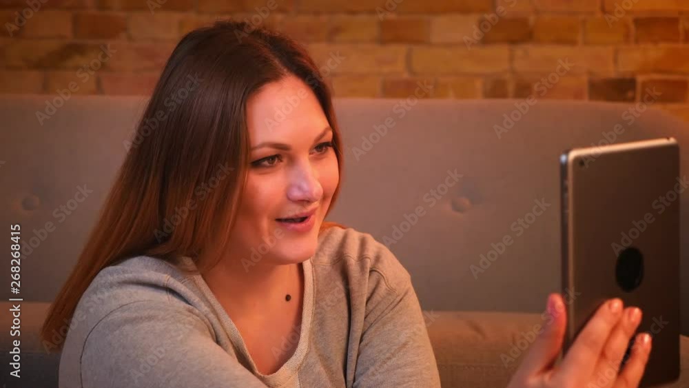 Close-up portrait of overweight joyful female model sits on floor talking in videochat on tablet in cozy home atmosphere. Close-up portrait of overweight joyful female model sits on floor talking in videochat on tablet in cozy home atmosphere.