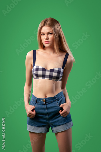 Portrait of a beautiful young  Caucasian woman 20 years old model in a black and white swimsuit and denim shorts with natural make-up posing on green isolated background © Виталий Сова