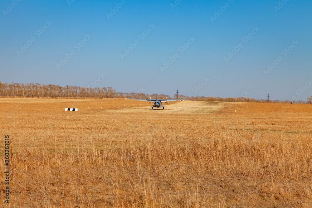 Obraz premium A small aircraft for the transport of passengers and paratroopers lands in a field on a landing strip with grass under a blue sky above the trees on a clear cloudless day. Air patrol.