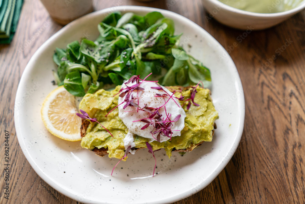 Morning in cafe, oak table. Healthy breakfast with wholemeal bread toast with avocado, poached egg with green salad. Green tea on the Background. Morning in cafe, oak table. Healthy breakfast with wholemeal bread toast with avocado, poached egg with green salad. Green tea on the Background.