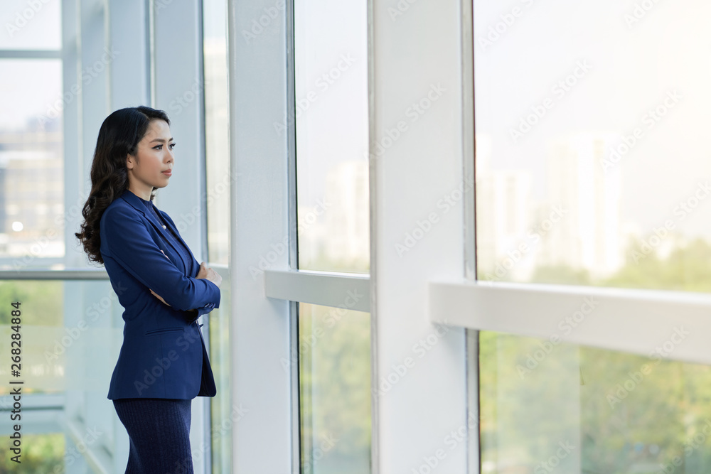 Young Asian businesswoman standing with arms crossed near the big window at office looking at view of the city and thinking about something Young Asian businesswoman standing with arms crossed near the big window at office looking at view of the city and thinking about something