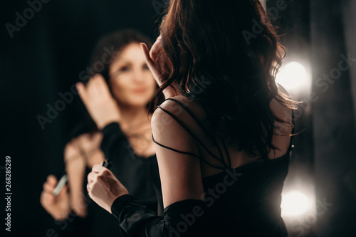 beautiful elegant brunette in a black silk dressing gown is standing in front of a tall make-up mirror and dressing herself up, paints eyelashes with mascara in a gray luxurious bathroom