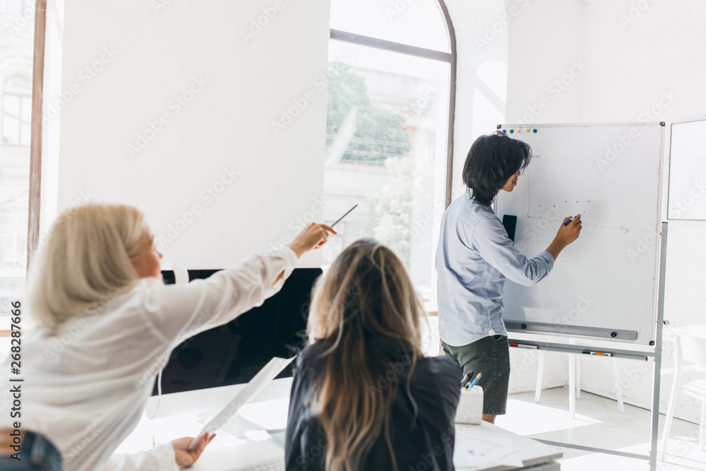 Obraz premium Businessman with black hair drawing infographic to explain new goals to colleagues. Indoor portrait from back of two ladies looking at asian guy beside flipchart.