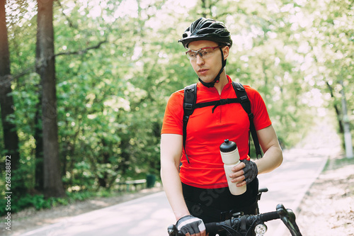 cyclist in a helmet and a red T-shirt is resting by the road, drinking water