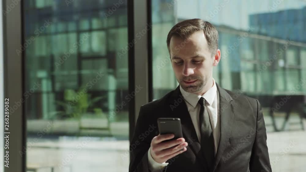 Panning shot of middle aged businessman in formal suit and tie reading and sending messages on cell phone standing in entrance hall of business center and looking away towards sun through glass wall Panning shot of middle aged businessman in formal suit and tie reading and sending messages on cell phone standing in entrance hall of business center and looking away towards sun through glass wall