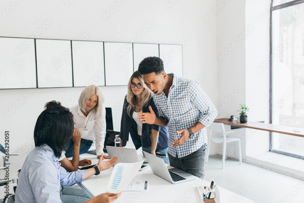 Obraz premium Angry black chief standing near table and waving hands. Indoor portrait of unhappy manager looking at laptop screen and telling something to asian programmer in blue shirt.