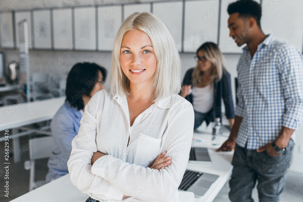 Blue-eyed business-woman in white blouse standing in confident pose with her international co-workers on background. Indoor portrait of asian and african employees with blonde lady on foreground. Blue-eyed business-woman in white blouse standing in confident pose with her international co-workers on background. Indoor portrait of asian and african employees with blonde lady on foreground.