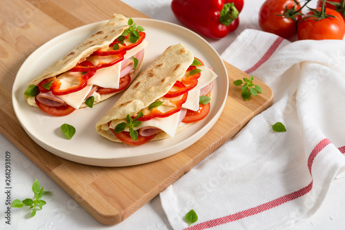 Italian piadina romagnola flatbread with red pepper, tomatoes, prosciutto ham, cheese and basil on the plate on white wooden background. Copy space