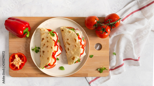 freshly baked italian piadina with cheese, tomato, ham, red pepper and basilio on a white plate on a white wooden table. Top view, flat lay. Copy space, banner for restaurant photo