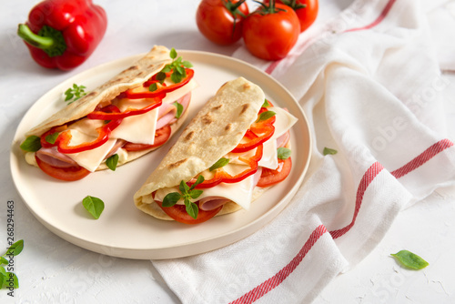 Italian piadina romagnola flatbread with red pepper, tomatoes, prosciutto ham, cheese and basil on the plate on white wooden background. Copy space