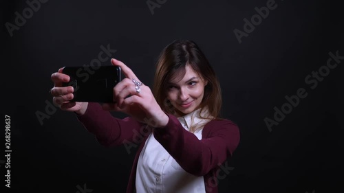 Portrait of young brunette businesswoman making beautiful selfie-photos on cellphone happily on black background. photo