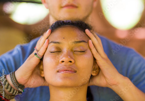 natural lifestyle portrait of young beautiful and relaxed Asian Balinese woman receiving a healing facial and head Thai massage by male therapist at traditional spa