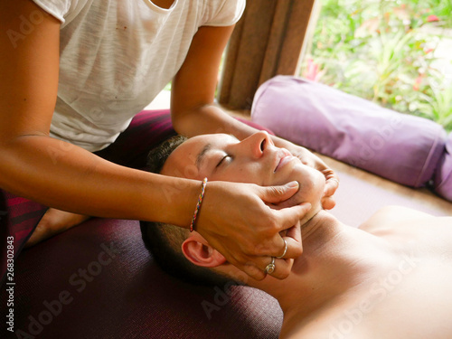 young beautiful and exotic Asian Thai therapist woman giving traditional head and facial Balinese massage to Caucasian man at alternative medicine healing spa photo