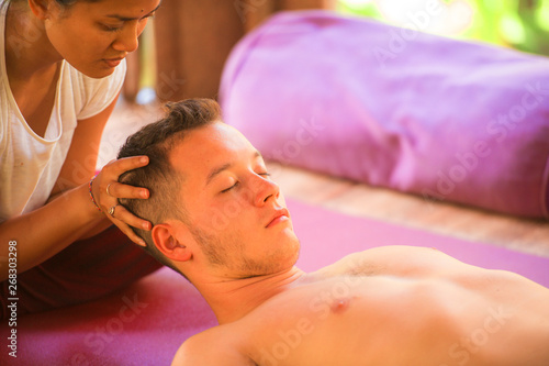 young beautiful and exotic Asian Thai therapist woman giving traditional head and facial Balinese massage to Caucasian man at alternative medicine healing spa