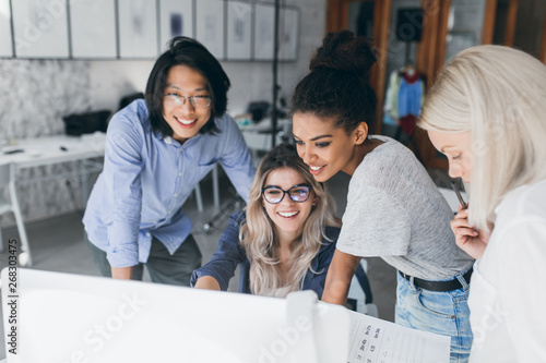 Obraz na plátně Long-haired blonde secretary in glasses laughing while looking at computer with asian programmer and african girl