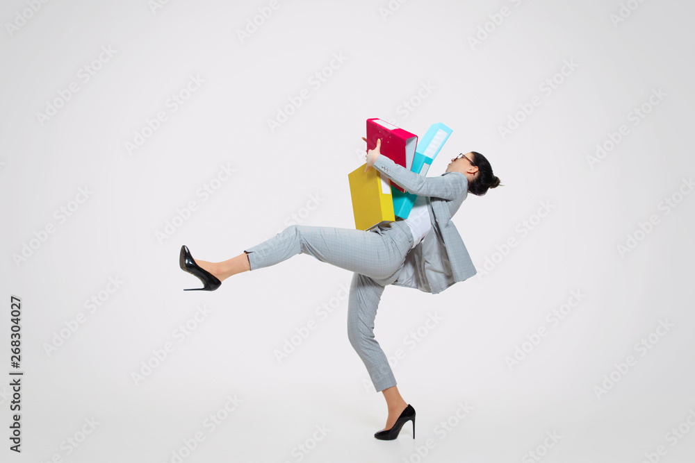 Fototapeta premium Young woman in suit getting a lot of work and losing concentration. Dropping folders with papers on the go, trying to catch that. Concept of office worker's troubles, business, problems and stress.