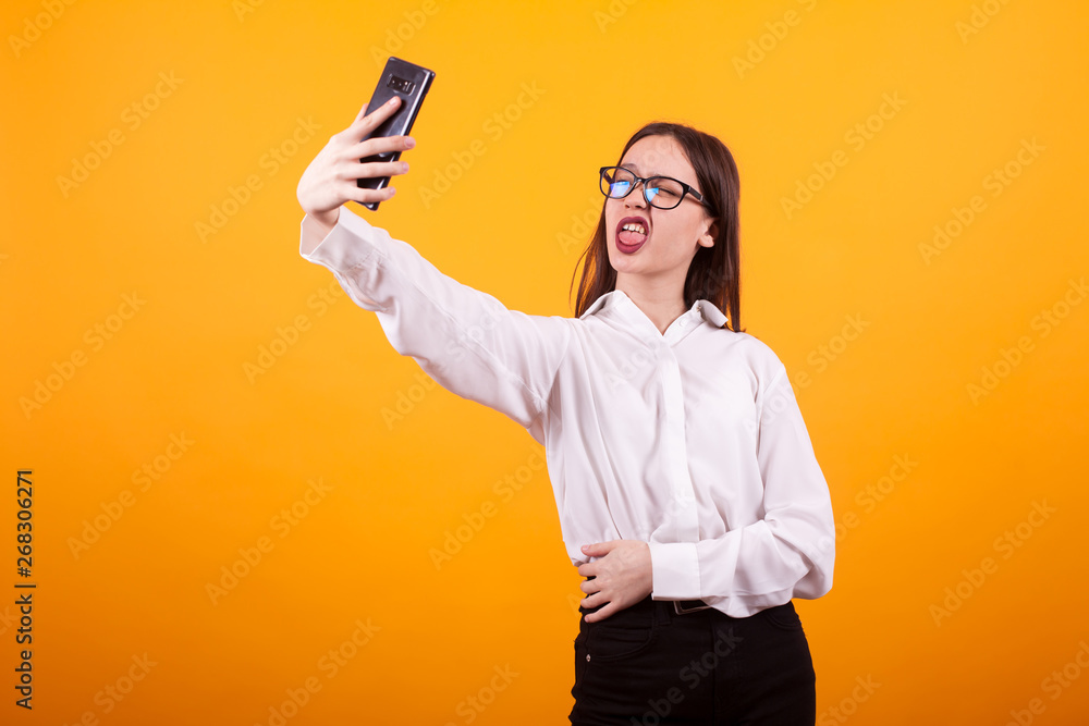 Pretty young teenage girl making silly face and taking a selfie in studio over yellow background