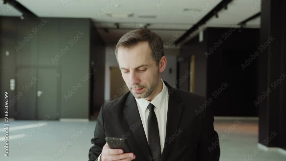 Dolly medium shot of middle aged businessman in black formal suit and necktie text messaging on cell phone while walking along office building corridor Dolly medium shot of middle aged businessman in black formal suit and necktie text messaging on cell phone while walking along office building corridor