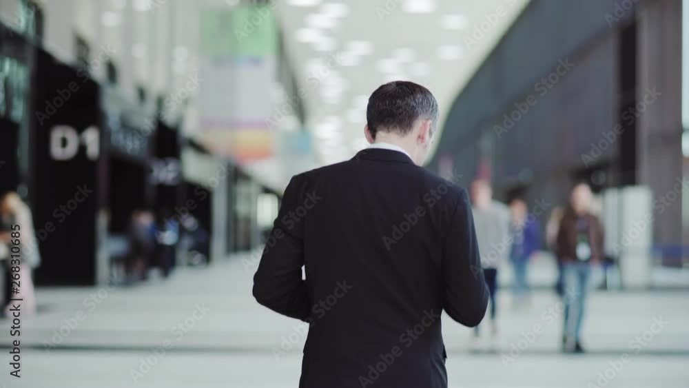 Follow rear view shot of businessman in suit walking along hall in business center, airport or shopping mall and using cell phone on move Follow rear view shot of businessman in suit walking along hall in business center, airport or shopping mall and using cell phone on move