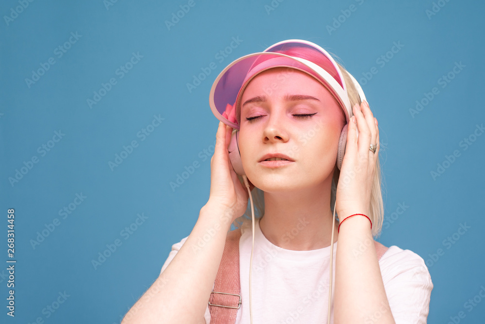 Closeup portrait of a girl in a white T-shirt and a pink cap, listens to music in her headphones with her eyes closed on a blue background. Isolated. Music concept. Closeup portrait of a girl in a white T-shirt and a pink cap, listens to music in her headphones with her eyes closed on a blue background. Isolated. Music concept.