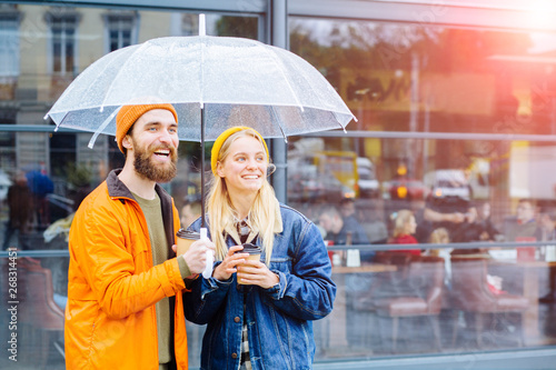 Canvas Print Positive bearded man with transparent umbrella and hiis girlfriend in bright wear complementary colors laughing looking aside in rainy weather urban city street on background