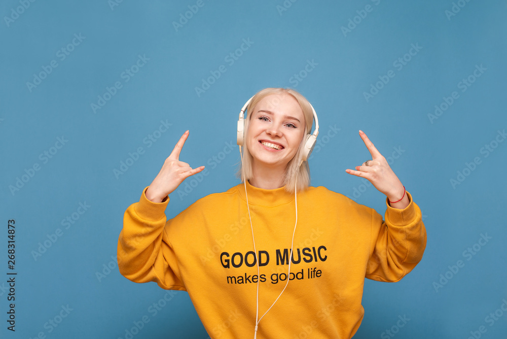 Happy blonde in an orange sweatshirt and headphones is on a blue background, listens to music and shows a sign of heavy metal, looks into the camera and smiles. Isolated Happy blonde in an orange sweatshirt and headphones is on a blue background, listens to music and shows a sign of heavy metal, looks into the camera and smiles. Isolated