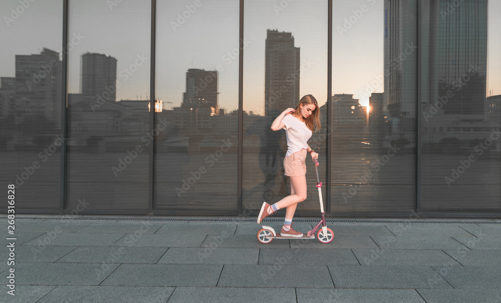Φωτογραφία Stylish blond girl in a white T-shirt with a pink kick scooter poses against the background of a glass shop window