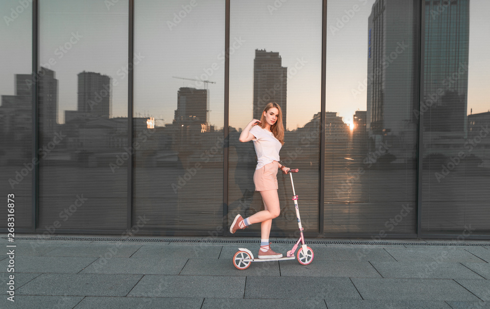 Φωτογραφία Portrait of attractive girl riding on pink scooter against background of dark glass wall, reflection of sun and city landscape against background