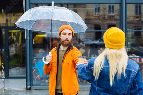 Hipster smiling beard man in orange hat and jacket drinks coffee to go under transparent umbrella meeting his blond girlfriend in blue denim jacket and yellow hat in rainy urban street on background. photo