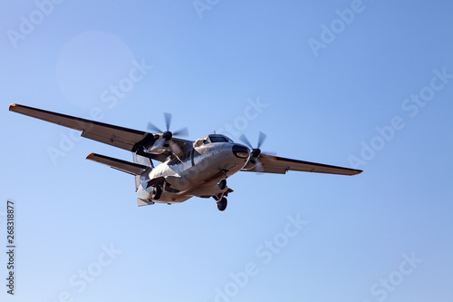 A small aircraft for the transport of passengers and paratroopers lands in a field on a landing strip with grass under a blue sky above the trees on a clear cloudless day. Air patrol.