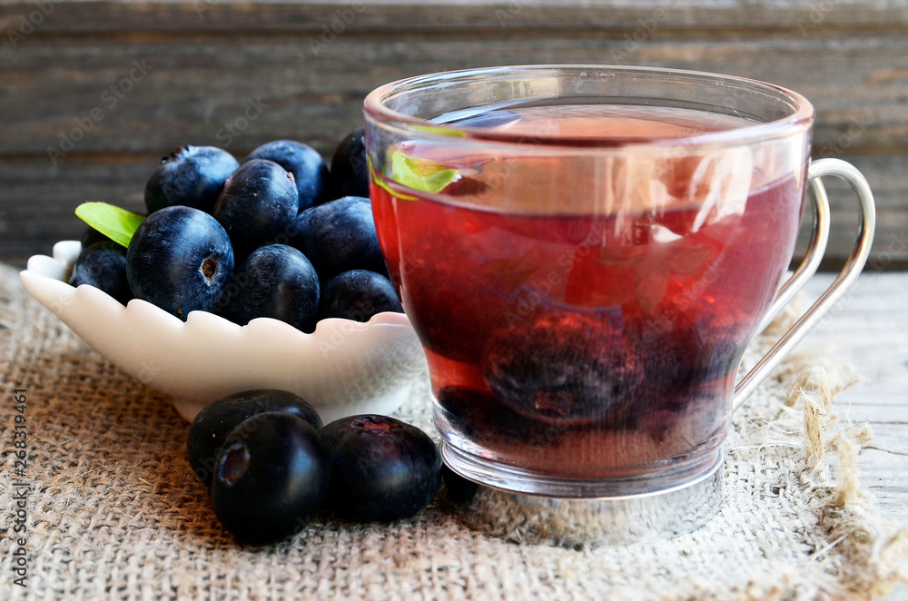 Blueberry tea in a glass cup and fresh blueberries on a burlap cloth on wooden background.Natural herbal summer beverage.Selective focus. Blueberry tea in a glass cup and fresh blueberries on a burlap cloth on wooden background.Natural herbal summer beverage.Selective focus.