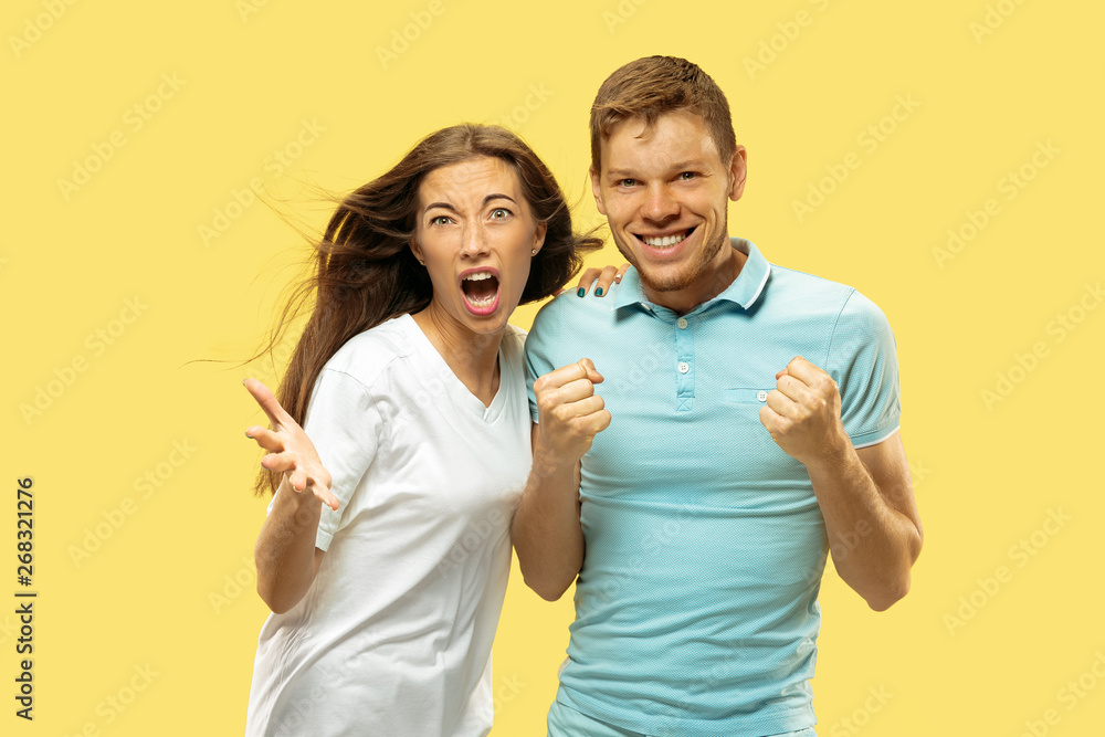 Fototapeta premium Beautiful young couple's half-length portrait on yellow studio background. Woman and man in shirts celebrating something and happy. Facial expression, human emotions concept. Trendy colors.
