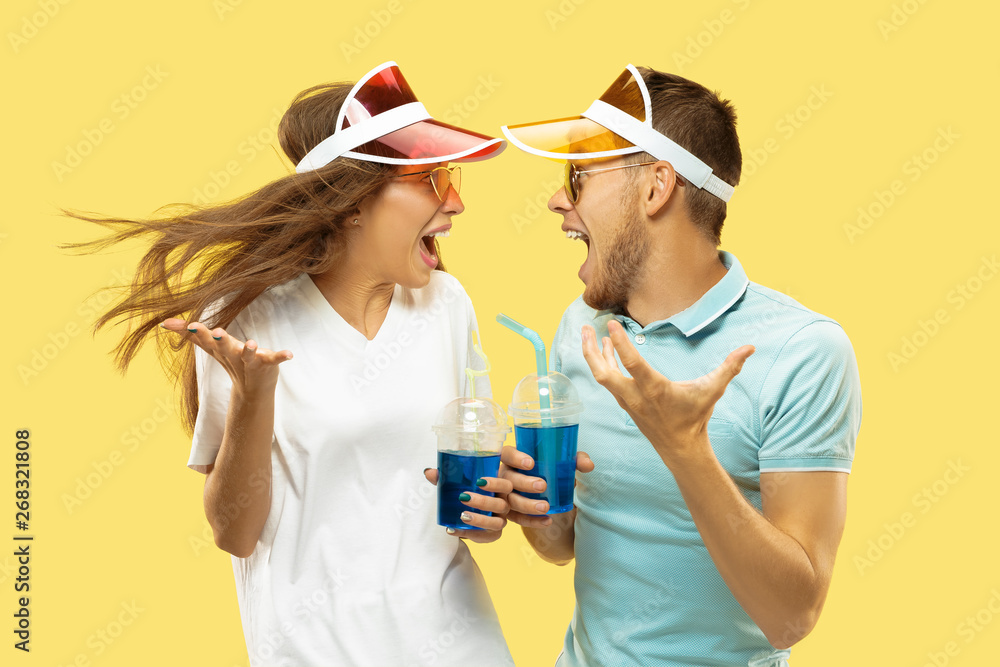 Beautiful young couple's half-length portrait isolated on yellow studio background. Woman and man standing with drinks in colorful caps. Facial expression, summer, weekend concept. Trendy colors. Beautiful young couple's half-length portrait isolated on yellow studio background. Woman and man standing with drinks in colorful caps. Facial expression, summer, weekend concept. Trendy colors.