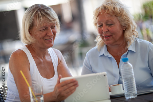 Two smiling senior women are sitting close to each other at a table of an outdoor cafe. They are both looking at the tablet that one of the women is holding in a hand. There are a bottle of water, a