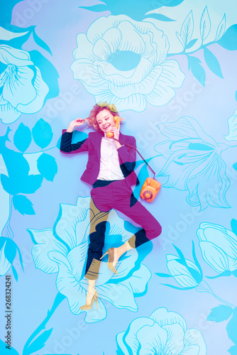 top view of businesswoman in suit and high-heeled shoes talking on telephone while lying on blue background with floral illustration