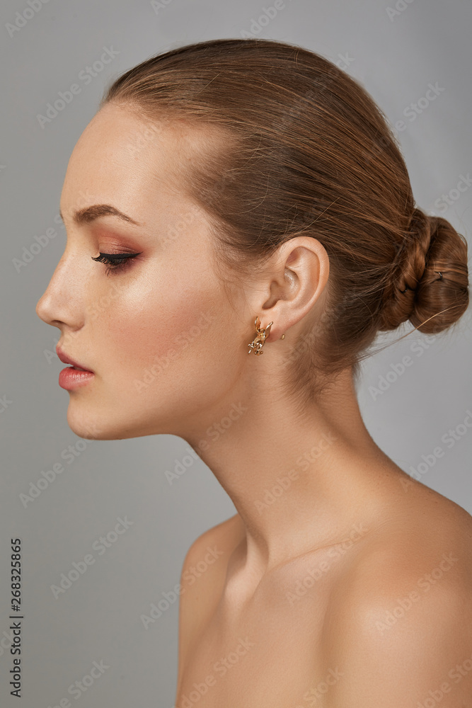 Cropped side view shot of woman with chestnut hair, tied back in a bun, with naked chest and fresh makeup, looking down. The girl with tanned skin is wearing golden earring in the shape of Pegasus. Cropped side view shot of woman with chestnut hair, tied back in a bun, with naked chest and fresh makeup, looking down. The girl with tanned skin is wearing golden earring in the shape of Pegasus.