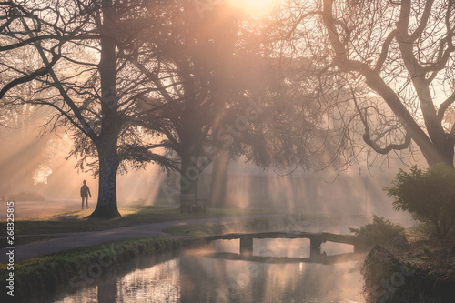 Cotswolds Lower Slaughter on a misty calm morning with sun shining through trees onto River Windrush