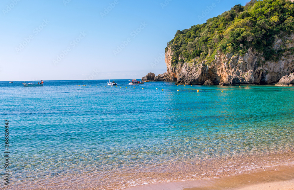 View of beautiful beach with clear blue water, golden sand, colorful boats and mountain on background. Summer landscape of Paleokastritsa  on Corfu Island, Greece and Ionian Sea. 