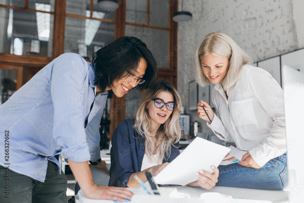 Fototapeta premium Beautiful blonde girl in glasses showing report to asian colleague which standing beside her table. Indoor portrait of cheerful international students analysing exam tasks.