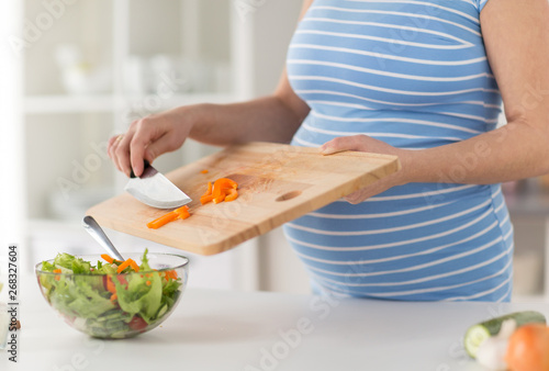 pregnancy, cooking food and healthy eating concept - close up of pregnant woman with kitchen knife adding chopped pepper from wooden cutting board to salad bowl at home