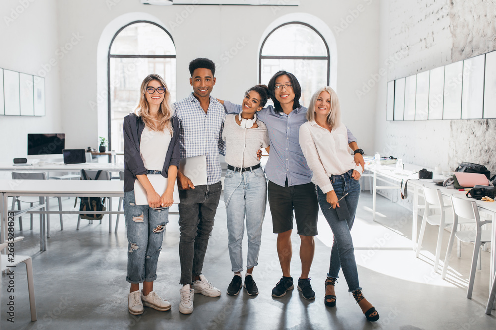 Fototapeta premium Full-length portrait of shy blonde girl in white sneakers holding laptop after seminar and stands beside african friend. Excited international students posing together after lecture in spacious hall.